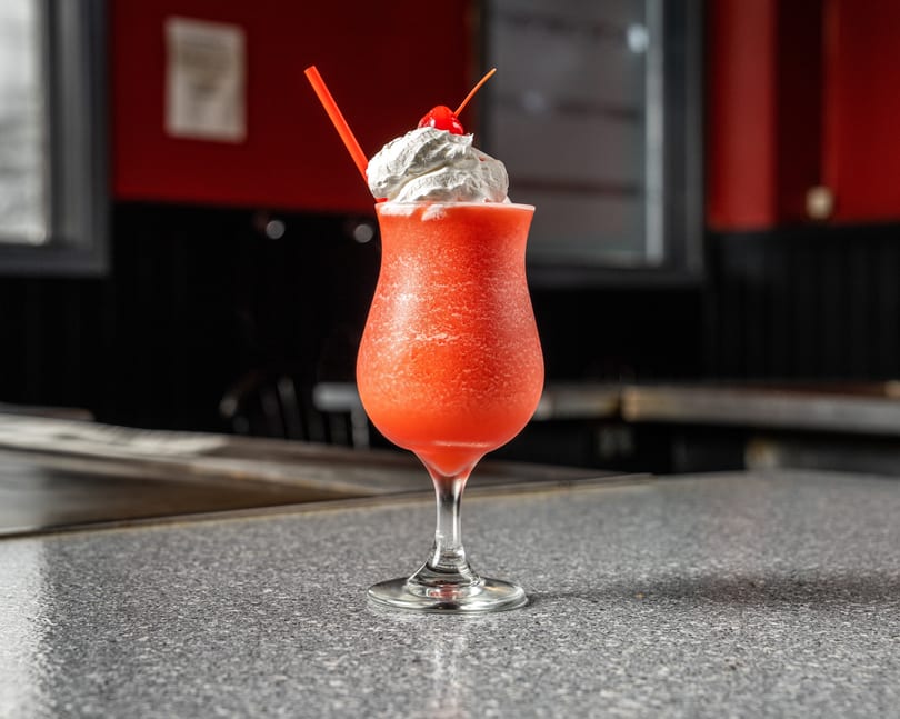 A vibrant red frozen drink with whipped cream, a cherry, and a straw on a speckled countertop.