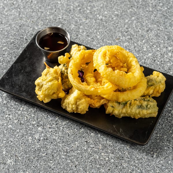 Golden-fried tempura vegetables and onion rings on a black rectangular plate with a side of dipping sauce.