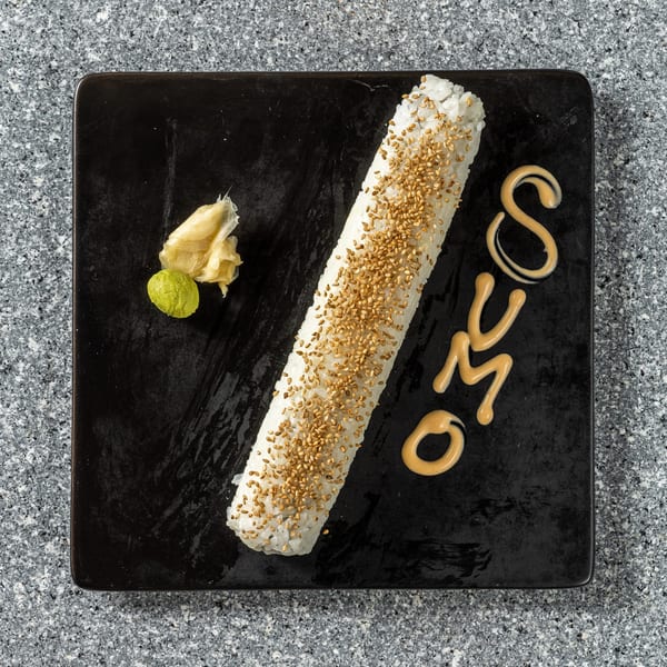 Long sushi roll with sesame seeds, wasabi, ginger, and "SUMO" written in sauce on a black plate.