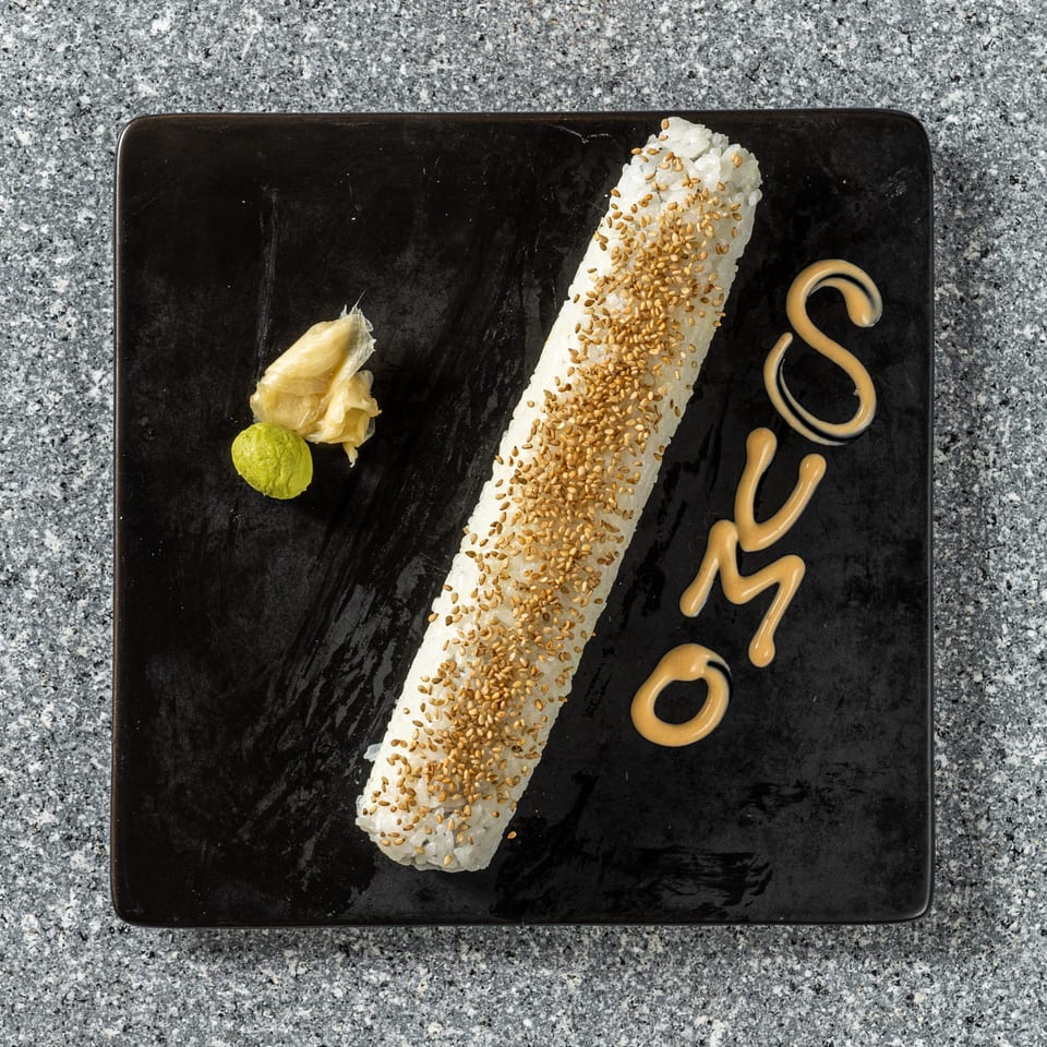 Long sushi roll with sesame seeds, wasabi, ginger, and "SUMO" written in sauce on a black plate.