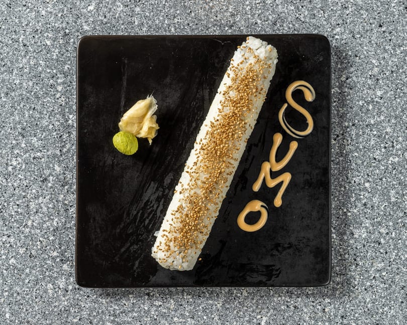 Long sushi roll with sesame seeds, wasabi, ginger, and "SUMO" written in sauce on a black plate.