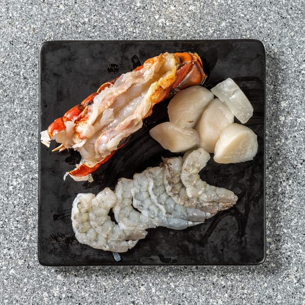 Raw lobster tail, scallops, and shrimp on a black plate, ready for cooking.