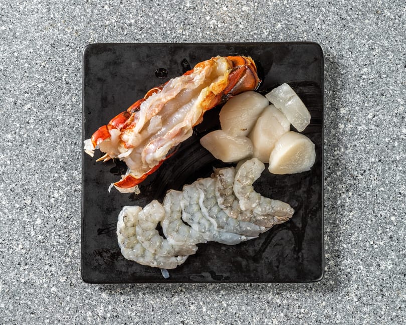 Raw lobster tail, scallops, and shrimp on a black plate, ready for cooking.