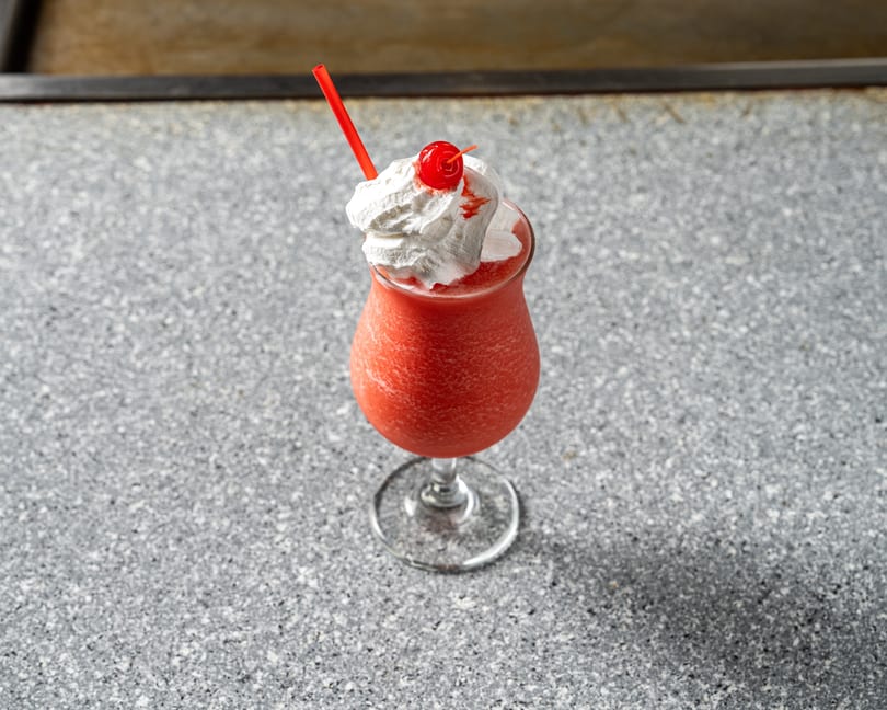 A red frozen drink with whipped cream, a cherry, and a red straw on a speckled gray surface.