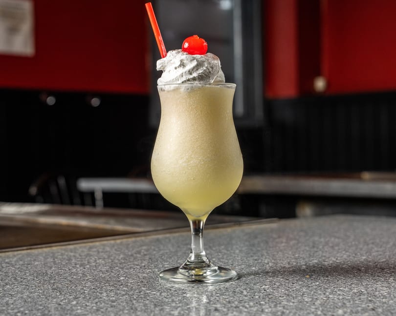 A frozen cocktail with whipped cream, a cherry, and a red straw sits on a speckled gray bar.