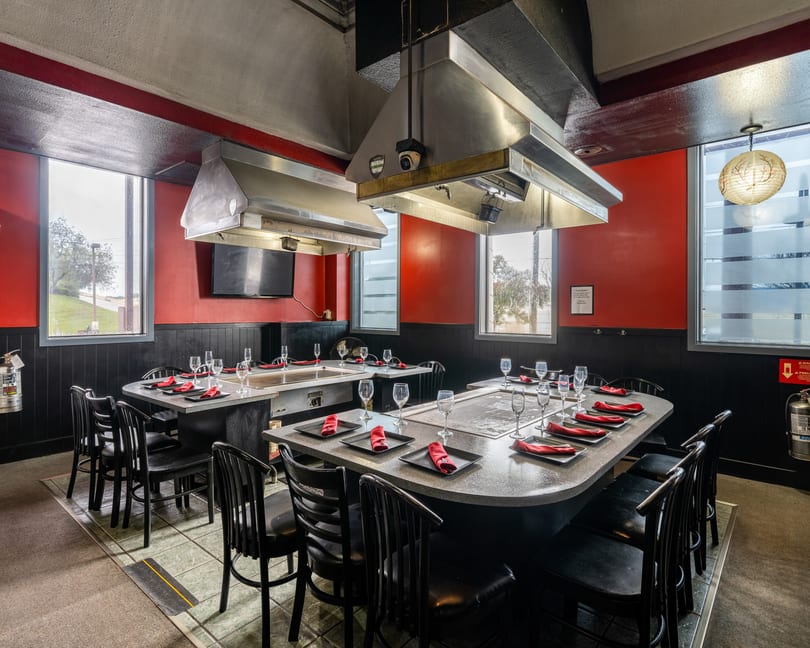Hibachi restaurant dining room with two large tables, red walls, black chairs, and cooking griddles.