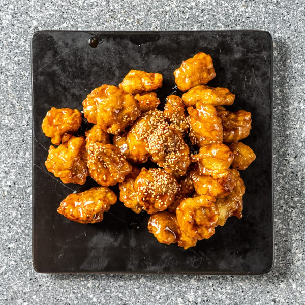 Sweet and sour chicken bites, glazed with sauce and sprinkled with sesame seeds on a black plate.