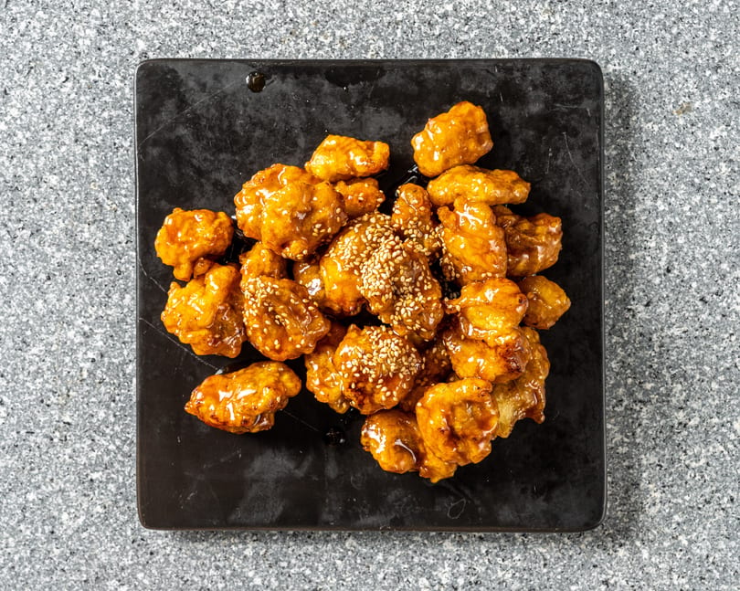 Sweet and sour chicken bites, glazed with sauce and sprinkled with sesame seeds on a black plate.