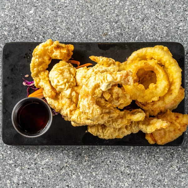 Crispy golden-brown tempura shrimp and onion rings with a side of dark dipping sauce on a black platter.