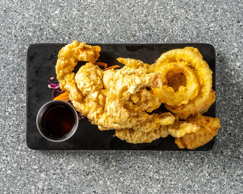 Crispy golden-brown tempura shrimp and onion rings with a side of dark dipping sauce on a black platter.