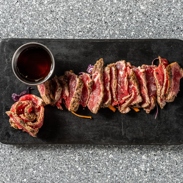 Seared beef tataki slices with a side of sauce and garnish on a black rectangular plate.
