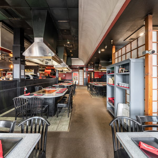 A long, narrow restaurant dining room with hibachi tables, black chairs, and red accents.
