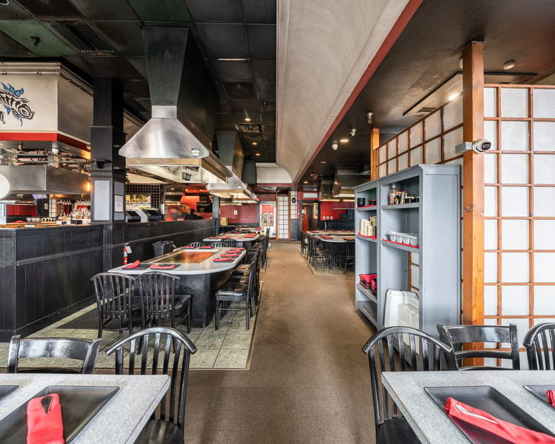 A long, narrow restaurant dining room with hibachi tables, black chairs, and red accents.
