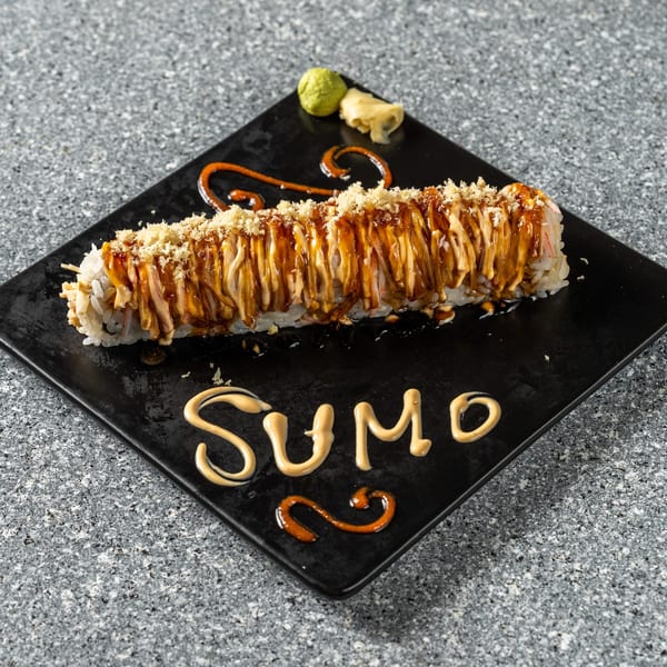 A long sushi roll with sauces and crispy toppings on a black plate, with "SUMO" written in sauce.