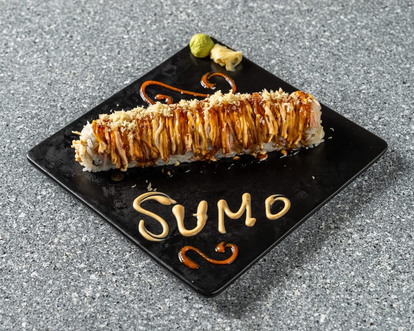 A long sushi roll with sauces and crispy toppings on a black plate, with "SUMO" written in sauce.
