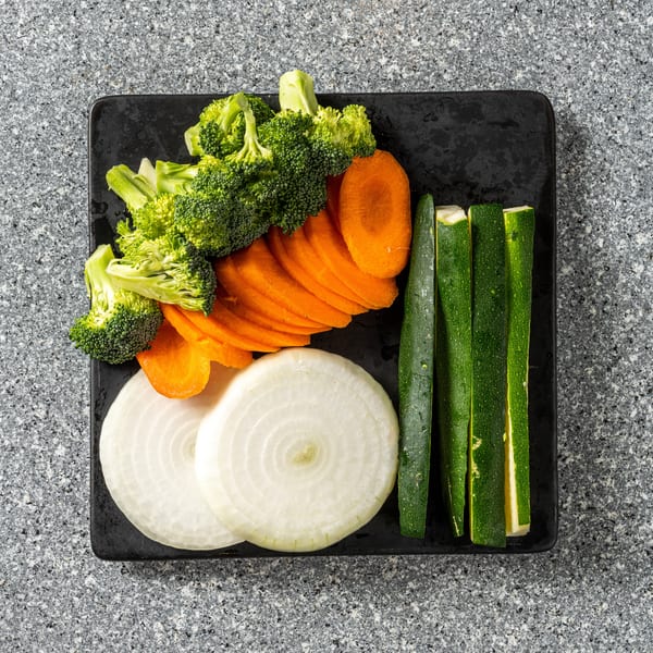 Top-down view of a black plate with sliced broccoli, carrots, onions, and zucchini on a gray counter.