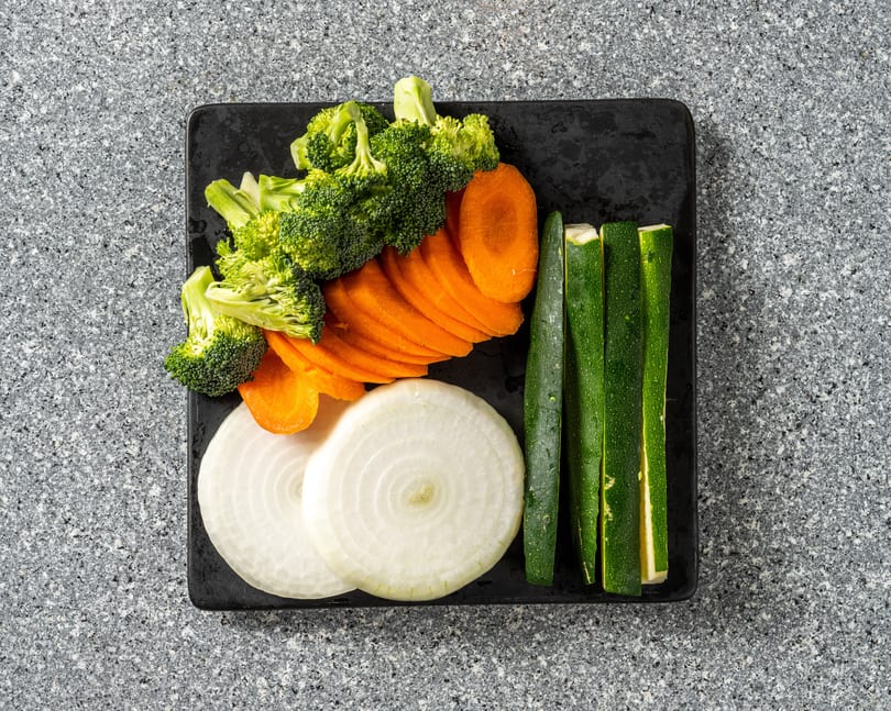 Top-down view of a black plate with sliced broccoli, carrots, onions, and zucchini on a gray counter.