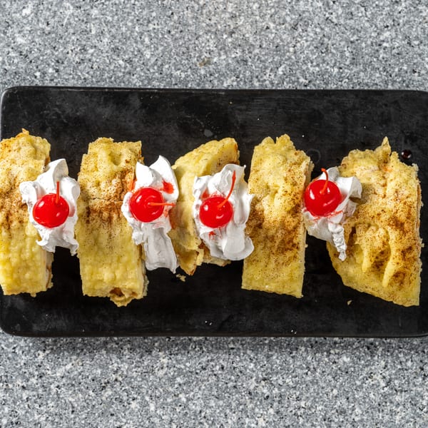 Five golden-brown dessert pieces, topped with whipped cream and a cherry, on a black rectangular plate.