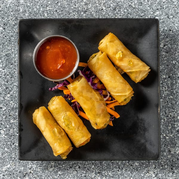 Five golden spring rolls with shredded carrots and purple cabbage, served with a side of red sauce.
