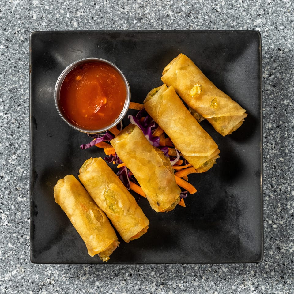 Five golden spring rolls with shredded carrots and purple cabbage, served with a side of red sauce.