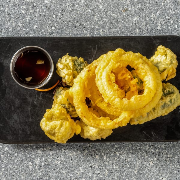 Golden tempura-fried vegetables and onion rings with a side of dark dipping sauce on a black slate.