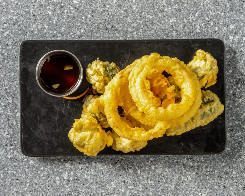 Golden tempura-fried vegetables and onion rings with a side of dark dipping sauce on a black slate.