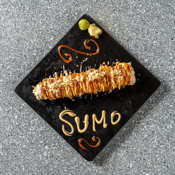 A long sushi roll with sauce and crispy toppings on a black plate, with "SUMO" written in sauce.