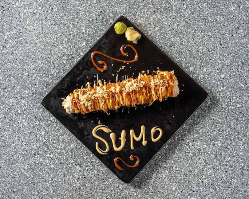 A long sushi roll with sauce and crispy toppings on a black plate, with "SUMO" written in sauce.