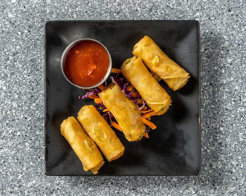 Five crispy spring rolls with shredded carrots and purple cabbage, served with a side of dipping sauce.