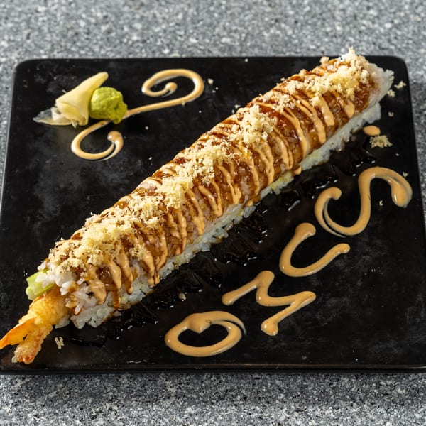 Long sushi roll with tempura shrimp, drizzled with sauces, served on a black plate with ginger and wasabi.