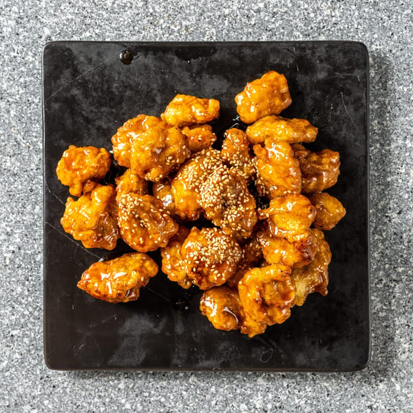 Sweet and sour chicken bites, glazed with sauce and sprinkled with sesame seeds on a black plate.