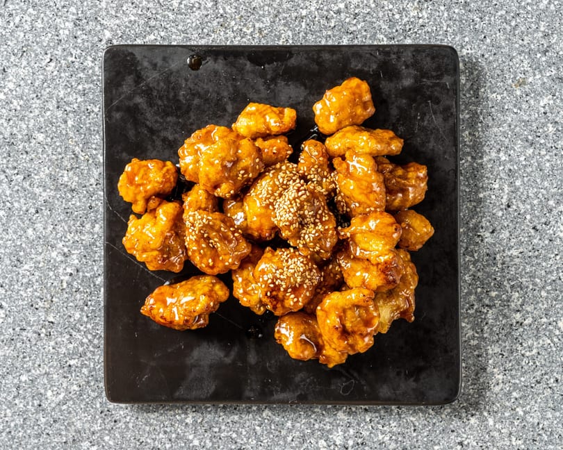 Sweet and sour chicken bites, glazed with sauce and sprinkled with sesame seeds on a black plate.