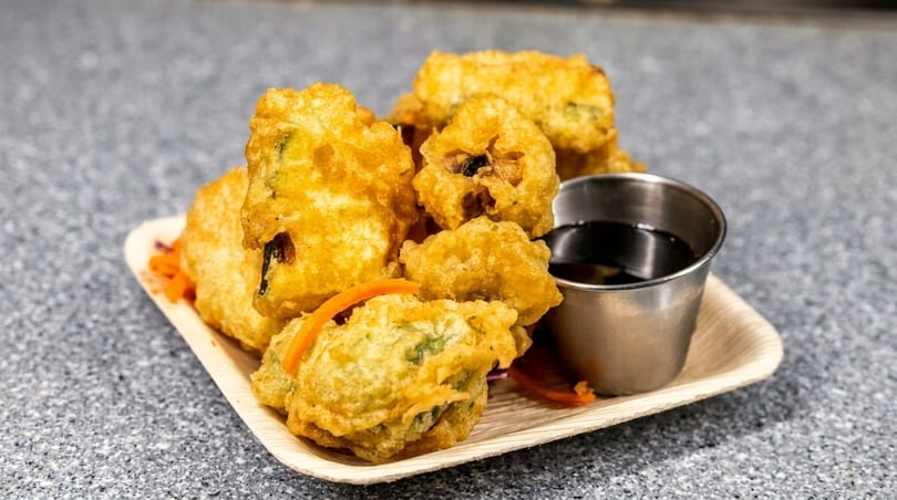 Golden fried tempura vegetables and shrimp with a side of dark dipping sauce on a wooden plate.