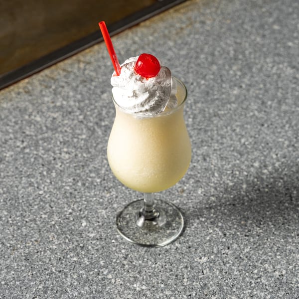 A yellow frozen drink with whipped cream, a red cherry, and a red straw on a speckled gray surface.