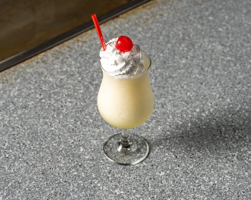 A yellow frozen drink with whipped cream, a red cherry, and a red straw on a speckled gray surface.
