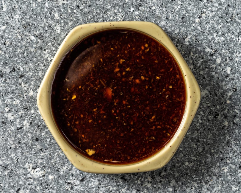 Overhead view of a hexagonal bowl filled with dark, spicy chili oil on a speckled gray surface.