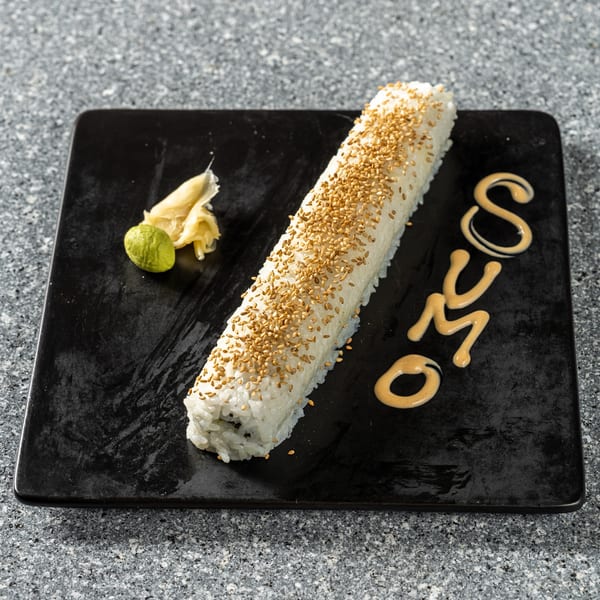 Long sushi roll topped with sesame seeds, with wasabi, ginger, and "SUMO" written in sauce.
