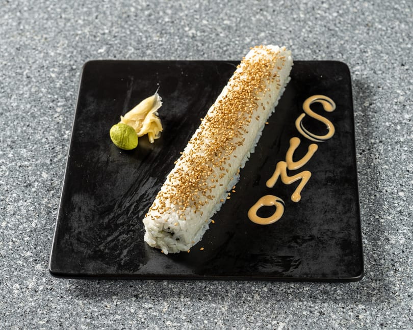 Long sushi roll topped with sesame seeds, with wasabi, ginger, and "SUMO" written in sauce.