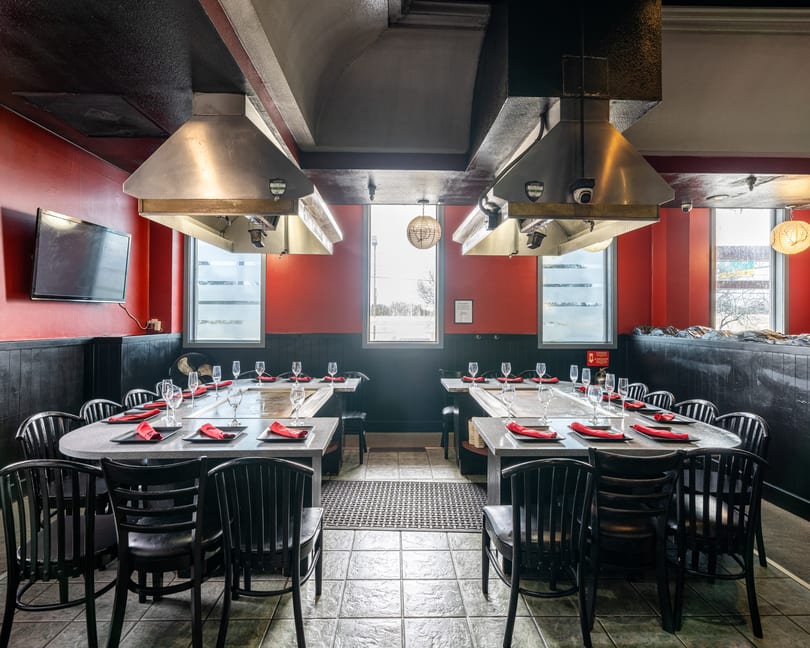 Empty hibachi dining room with red walls, black chairs, and long tables set with red napkins.