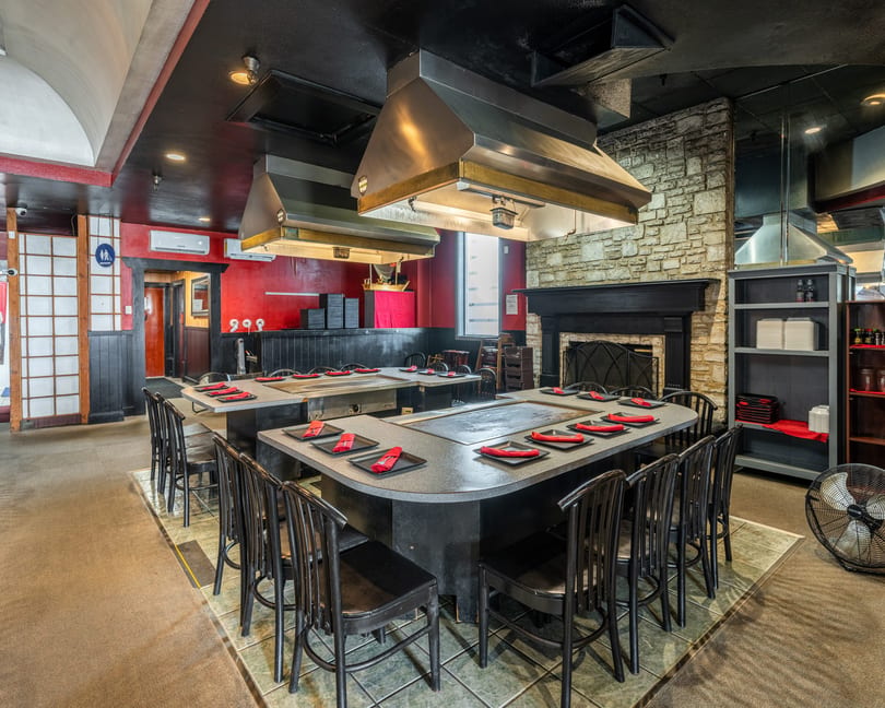 Empty hibachi restaurant dining room with red walls, black chairs, and cooking stations with red napkins.