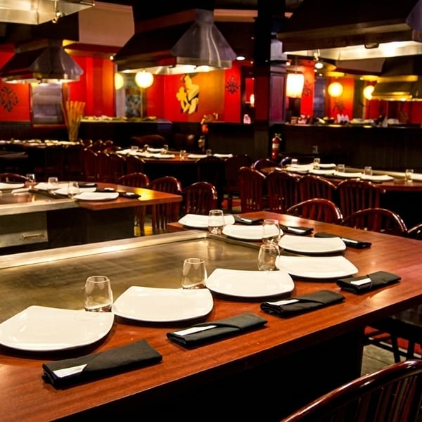 A dimly lit Japanese hibachi restaurant with multiple tables, each set with white plates and glasses.