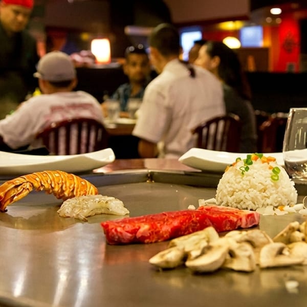 Teppanyaki grill with raw lobster, shrimp, steak, mushrooms, and rice, with diners in background.