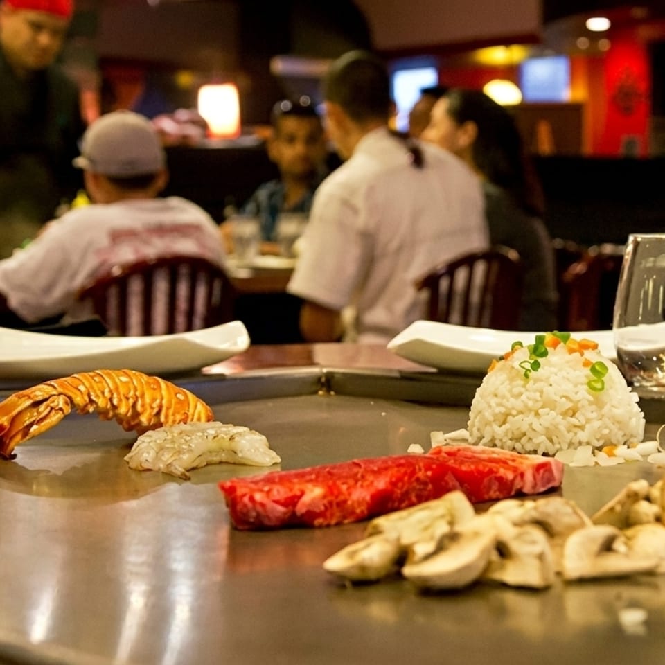 Teppanyaki grill with raw lobster, shrimp, steak, mushrooms, and rice, with diners in background.