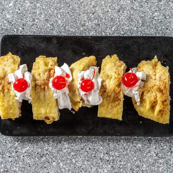Five golden-brown dessert pieces, topped with whipped cream and a cherry, on a black rectangular plate.