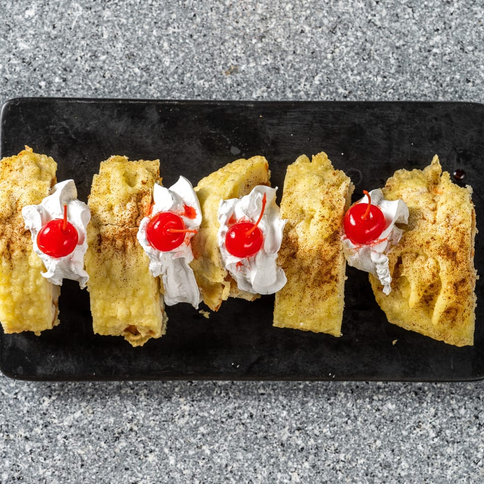 Five golden-brown dessert pieces, topped with whipped cream and a cherry, on a black rectangular plate.