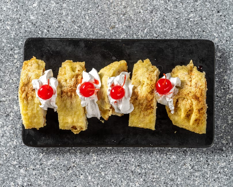 Five golden-brown dessert pieces, topped with whipped cream and a cherry, on a black rectangular plate.