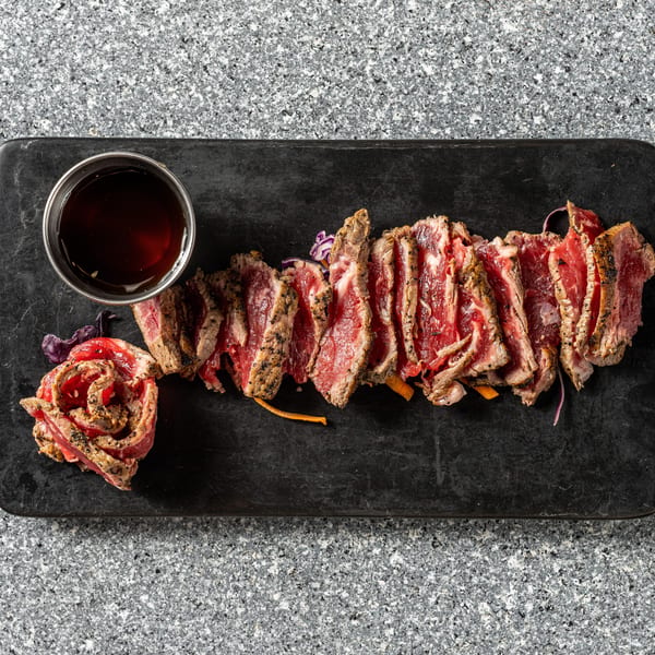 Seared beef tataki slices with sauce and garnishes on a dark rectangular plate.